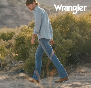 Man loopt op een zandpad in een woestijnlandschap, draagt lichtblauwe denim jeans, een denim overhemd en bruine laarzen, Wrangler-logo rechtsboven.