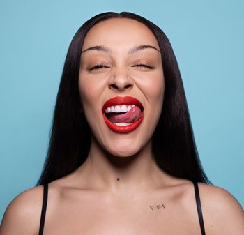Femme aux longs cheveux bruns et lèvres rouges vives se léchant les lèvres sur un fond bleu clair, portant un haut à bretelles noir.