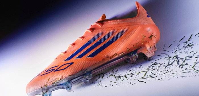 Chaussure de football orange avec un dessus texturé, des accents bleus et une semelle transparente. Détails d'herbe et de terre présents sur la semelle.