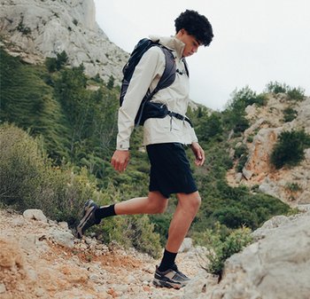 Une jeune personne portant un sac à dos descend un sentier rocheux entouré de buissons verts et de terrain montagneux.
