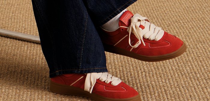 Sneakers rossi con lacci bianchi, realizzati in suede e materiale sintetico, con strisce laterali e suola in gomma marrone testurizzata, abbinati a pantaloni in denim.