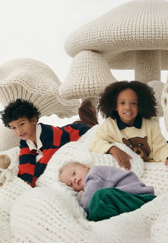 Τρία παιδιά κάθονται χαλαρά σε μια μεγάλη, χοντρή πλεκτή κουβέρτα, περιτριγυρισμένα από υπερμεγέθεις, λευκές, πλεκτές δομές σε σχήμα μανιταριού.