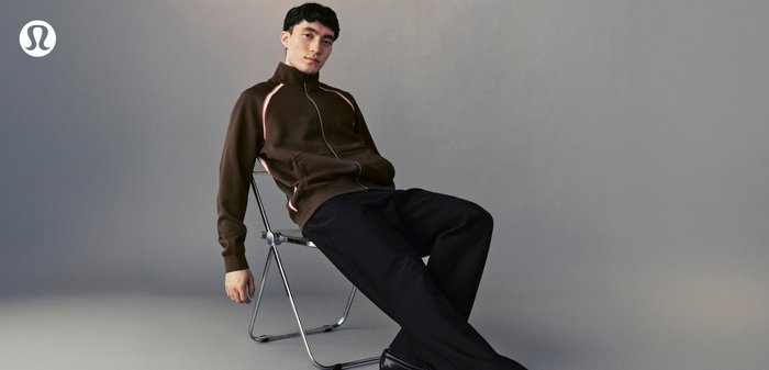 Junger Mann in brauner Jacke mit Reißverschluss und schwarzen Hosen lehnt sich auf einem Klappstuhl vor einem schlichten grauen Hintergrund zurück.