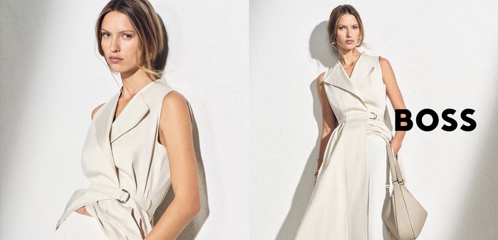 Woman with light brown hair wearing a sleeveless beige blazer dress, posing against a white wall with the word "BOSS" in bold black letters.