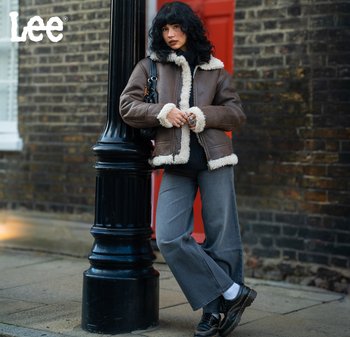 Kvinna i brun lammullsjacka och grå jeans som lutar sig mot en svart gatlykta på en stadsgata med tegelväggar.