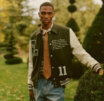 Veste de varsity noire et blanche avec "11" sur la poitrine gauche, portée par-dessus une chemise à carreaux et une cravate à motifs. Un pantalon en denim décontracté complète l'ensemble.