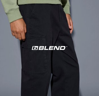 La mano sinistra poggia vicino alla patta della tasca dei pantaloni cargo neri, indossati con una maglietta a maniche lunghe verde chiaro, su uno sfondo grigio.