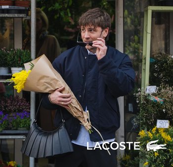Hombre con chaqueta azul marino sostiene un ramo de flores amarillas y un bolso negro frente a una floristería, ajustándose las gafas de sol.