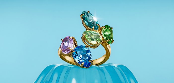 Bague en or avec cinq grosses pierres précieuses de formes différentes en bleu, violet, vert clair et vert, présentée sur une surface bleue texturée.