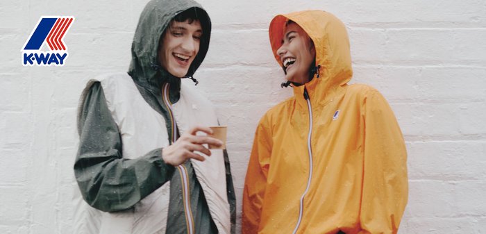 Zwei Personen lachen im Freien, eine in einer grauen und weißen Kapuzenjacke hält einen Becher, die andere in einer orangen Kapuzenjacke, vor einer weißen Wand.