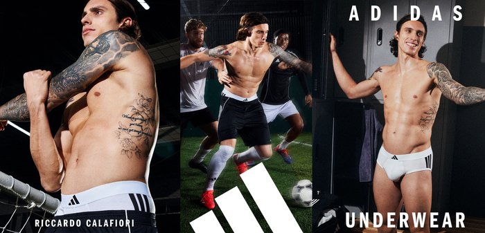 Slip da uomo Adidas, con un cinturino bianco decorato da tre strisce nere, pantaloncini neri, vestibilità atletica e tatuaggi visibili sul modello.