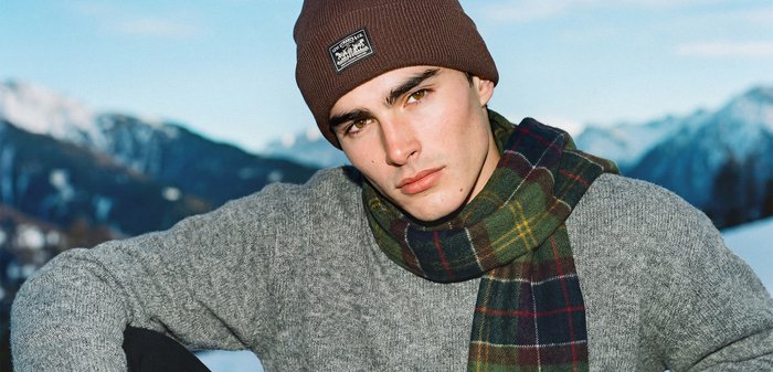 Bonnet tricoté marron, pull gris texturé et écharpe à carreaux verts avec des accents rouges et jaunes, sur fond de montagnes.