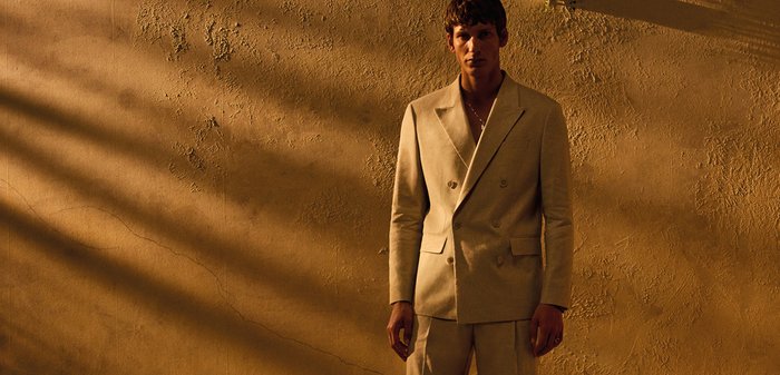 Homme debout pieds nus contre un mur texturé, portant un costume beige à double boutonnage sous une lumière chaude et basse du soleil.