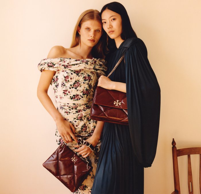 Due donne che tengono borse di design. Una indossa un vestito a spalle scoperte con stampa floreale; l'altra, un vestito scuro plissettato con una borsa a tracolla in catena. Entrambe le borse sono di colore marrone scuro.