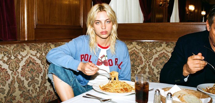 Jeune personne aux cheveux blonds portant un sweat-shirt bleu, mangeant des spaghettis à une table de restaurant avec une boisson et un panier à pain à proximité.
