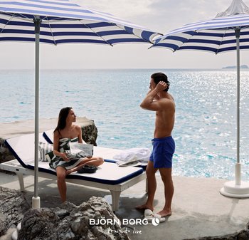 Kvinna sitter på en solstol insvept i en handduk och läser en tidning, man i blå badshorts står bredvid henne och fixar håret vid havet under randiga parasoller.