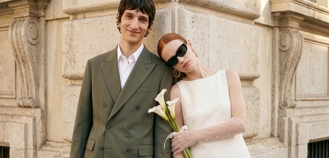 Hombre con traje verde sonríe mientras mujer con vestido blanco y gafas de sol se apoya en él, sosteniendo lirios cala blancos frente a un fondo de edificio de piedra.