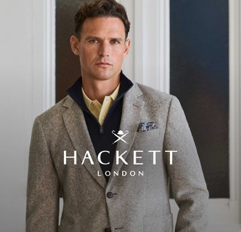 Homme portant un blazer gris clair par-dessus un sweat à capuche bleu marine zippé et un polo crème, debout à l'intérieur contre un mur blanc et bleu foncé.