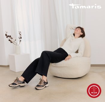 Cream-colored, textured bean bag chair; model in light sweater and wide-leg black pants, wearing patterned sneakers. Natural light background.