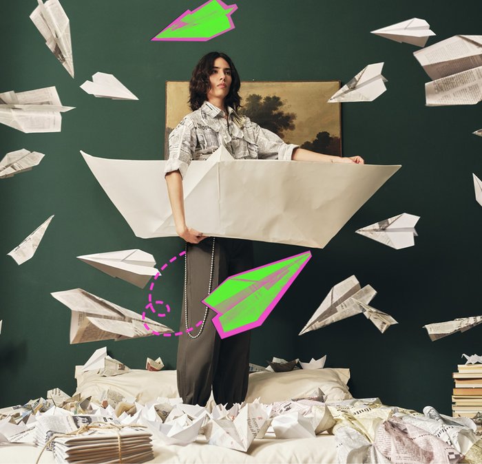 Person holding large paper boat surrounded by numerous paper planes and boats in a room with dark green walls and books stacked nearby.