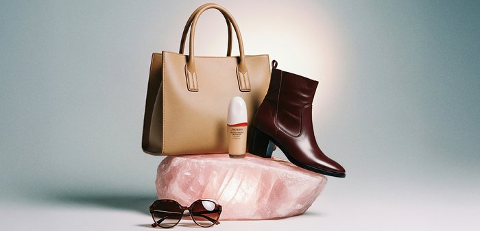 Beigetote, brauner Leder-Stiefelette, Flasche mit Foundation und Schildpattsonnenbrille, arrangiert auf und um einen großen rosa Kristall.
