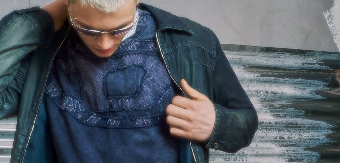 Ein junger Mann mit kurzem blondem Haar richtet seine dunkle Jacke, trägt ein gemustertes dunkelblaues Hemd, hellblaue Jeans und einen schwarzen Gürtel mit silberner Schnalle.