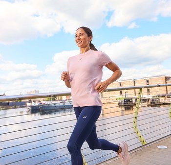Lichtroze sport-T-shirt met korte mouwen, gecombineerd met marineblauwe legging en lichtroze hardloopschoenen. Vrouw joggend langs een reling aan het water.