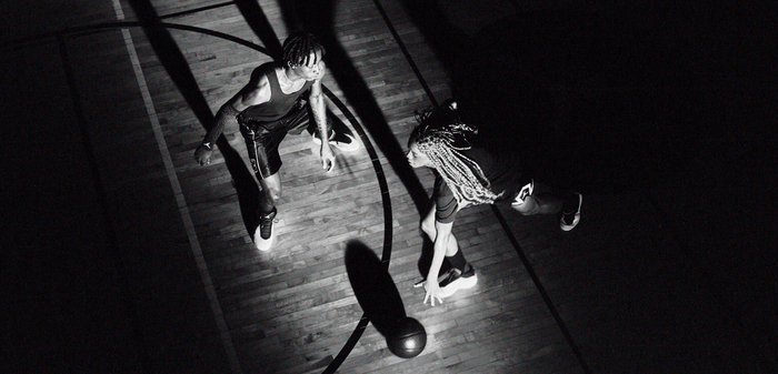 Deux joueurs de basket-ball, l’un dribblant et l’autre en défense, se font face sur un terrain éclairé de manière spectaculaire par un éclairage zénithal.