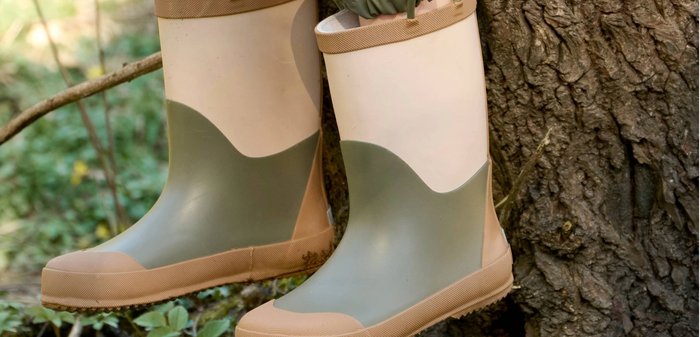 Kind trägt grüne und beige Gummistiefel, sitzt draußen mit baumelnden Füßen neben einem Baumstamm in natürlicher Umgebung.