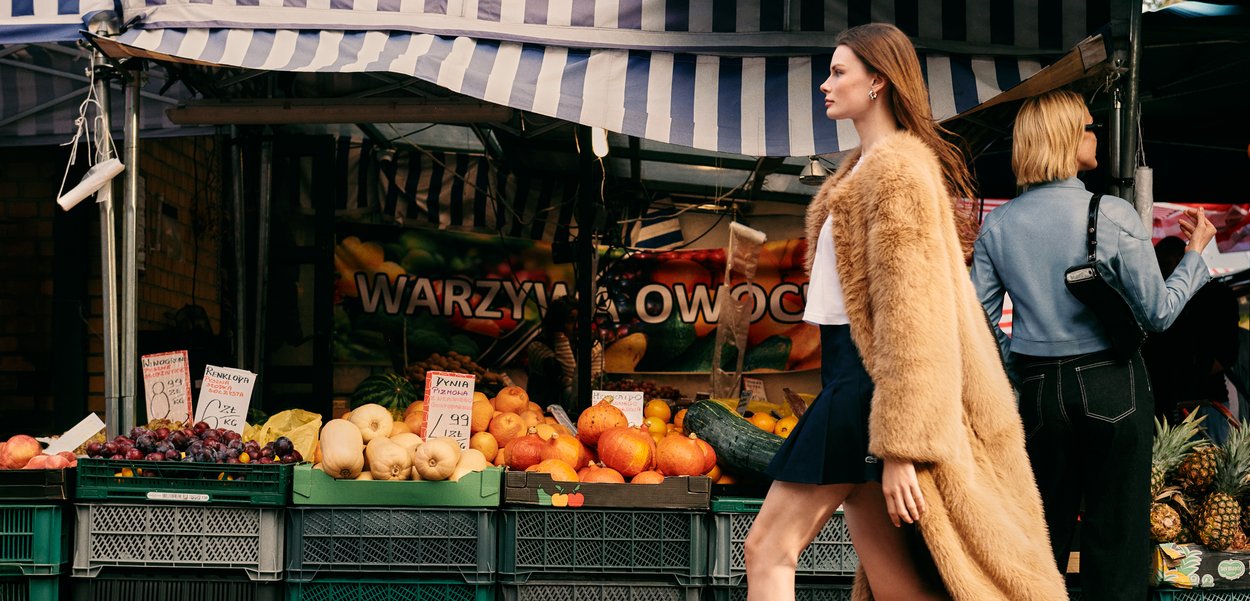Futrzana beżowa kurtka, biała bluzka i ciemna spódnica noszone przez kobietę przechodzącą obok rynku, gdzie wyeksponowane są kolorowe owoce i warzywa w skrzyniach.