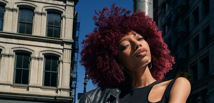 Giacca da biker nera in pelle appoggiata su un top senza maniche nero con un taglio. Capelli ricci color borgogna. Sfondo urbano con edifici.