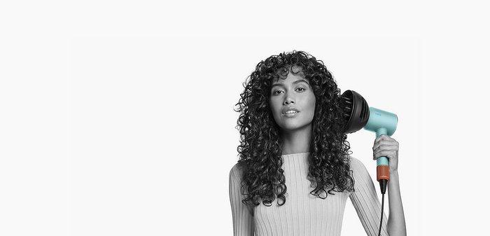 Een turquoise haardroger met een zwarte diffuseropzetstuk, vastgehouden door een persoon met krullend haar, tegen een witte achtergrond.