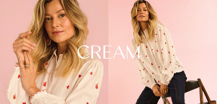 Blonde woman wearing cream blouse with red heart patterns, posing against a pink background with “CREAM” text overlay.