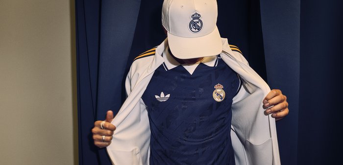 Άτομο που φοράει καπέλο της Ρεάλ Μαδρίτης και μπλε φανέλα Adidas της Ρεάλ Μαδρίτης, αποκαλύπτοντας τη φανέλα κάτω από ένα ανοιχτό λευκό μπουφάν.