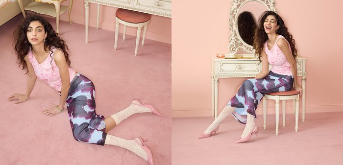 Jeune femme aux cheveux bouclés portant un haut rose, un pantalon à motifs et des chaussettes avec des talons, posant sur un sol rose et assise sur un tabouret vintage près d'une coiffeuse.