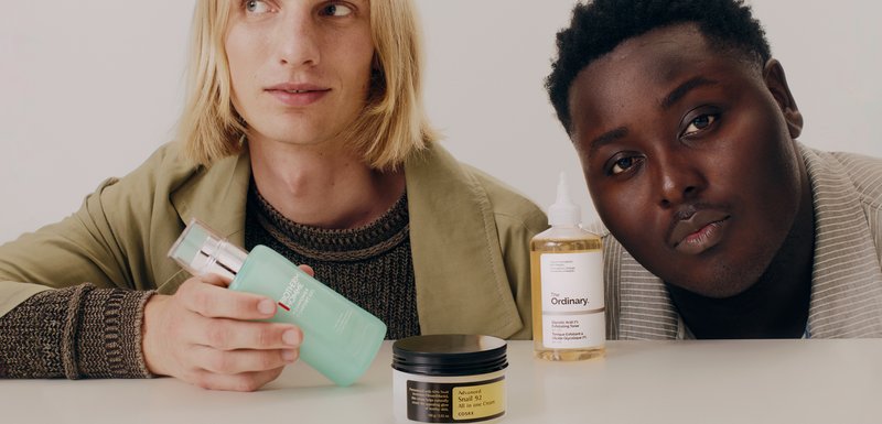 Trois produits de soin de la peau sont exposés sur une surface blanche : un gel nettoyant vert clair, un exfoliant liquide transparent et une crème en pot noir avec des étiquettes dorées.