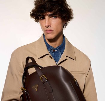 Zaino in pelle marrone con dettaglio in oro, caratterizzato da un design strutturato e una cerniera prominente. Il modello indossa una giacca beige e una camicia di jeans.