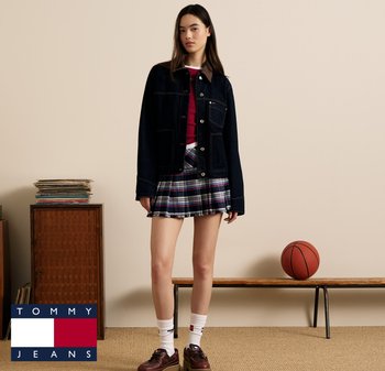 Veste en denim avec col marron, chemise rouge et jupe mini à carreaux. Chaussures marron, chaussettes blanches. Un ballon de basket et des meubles en bois en arrière-plan.