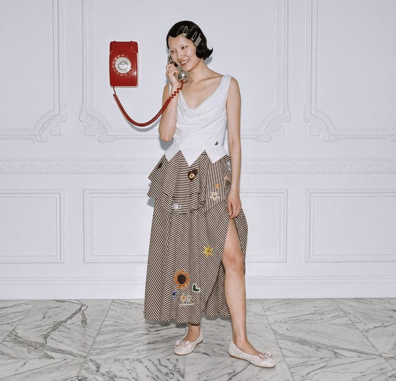 Femeie zâmbitoare ținând un receptor telefonic roșu cu disc, purtând un top alb fără mâneci și o fustă lungă cu dungi și modele brodate.