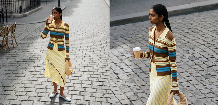 Femme aux cheveux tressés portant un pull rayé et une jupe jaune, tenant une tasse de café et un sac tissé, debout dans une rue pavée.