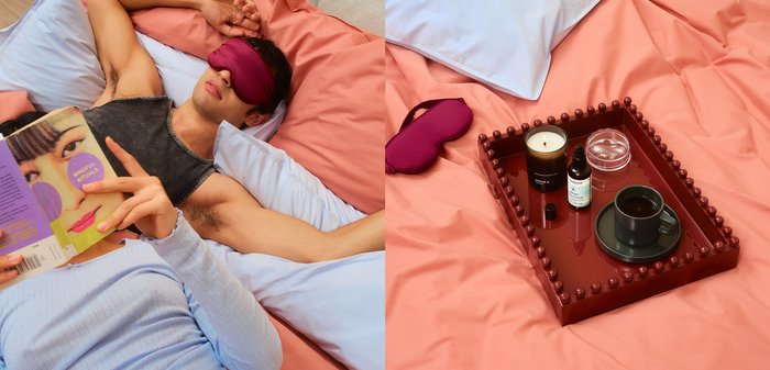 Une personne lit un livre sur un lit avec des draps roses et bleus. Un plateau contient une bougie, une huile essentielle, un récipient en verre et une tasse de café.