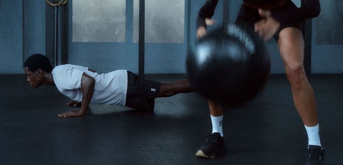 Deux personnes font des exercices dans une salle de sport ; l'une fait des pompes tandis que l'autre balance un gros ballon médicinal.