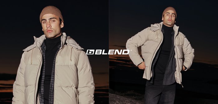 Mann trägt beige Daunenjacke, schwarzen Rollkragenpullover, kariertes Hemd und braune Mütze, steht draußen in der Dämmerung und posiert selbstbewusst.