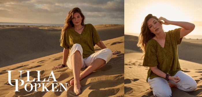Woman kneeling in sand dunes at sunset, wearing a textured olive green top and white pants, adjusting hair with one hand.