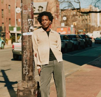 Jong persoon met afro-kapsel leunt tegen een graffiti-bedekte lantaarnpaal op een zonnige stedelijke straat met geparkeerde auto's.