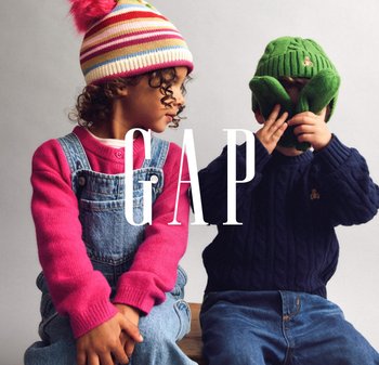 To børn sidder side om side. Den ene har en lyserød sweater og denim-odeskørt; den anden har en grøn strikket hat og en mørkeblå kabelstrikket sweater.