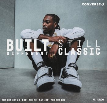 Hombre con cabello trenzado sentado contra una pared gris, vistiendo zapatillas Converse Chuck Taylor blancas, camisa blanca, pantalones negros y joyas doradas.