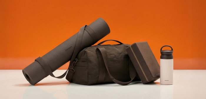 Gerollte Yogamatte, Sporttasche, Yoga-Block und Wasserflasche auf weißer Oberfläche vor orangefarbenem Hintergrund angeordnet.