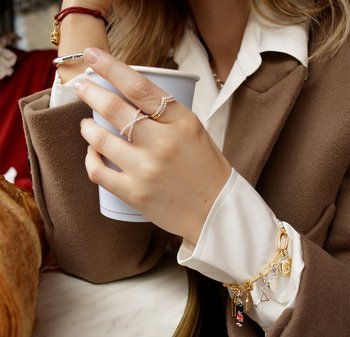 Gold and silver bangles and charm bracelet adorn a hand holding a blue coffee cup. A delicate twisted ring with clear stones is on a finger.