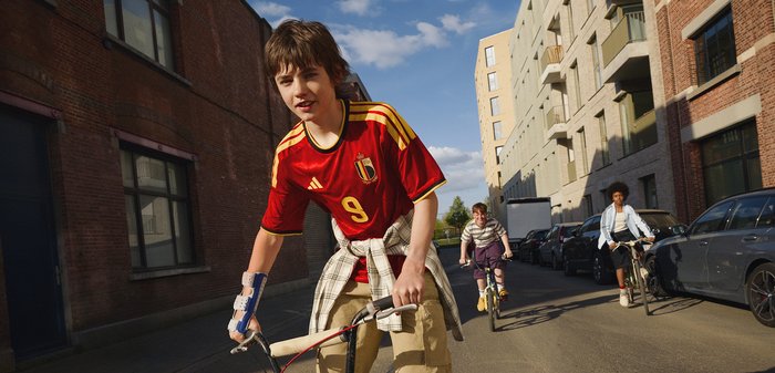 Trois garçons font du vélo dans une rue ensoleillée en ville, l’un porte un maillot rouge de football belge avec le numéro 9 et une attelle au poignet.